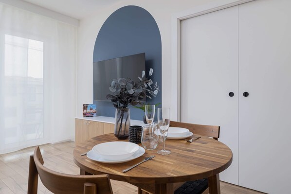 Un comedor con mesa de madera dispuesta para cuatro, un jarrón con flores y un televisor de pantalla plana montado en la pared.