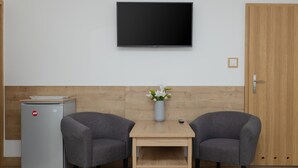 Una habitación con una televisión de pantalla plana montada en la pared, un refrigerador pequeño, dos sillones grises, una mesa de madera con un florero y una puerta de madera.