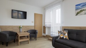 Una sala de estar moderna con un sofá, dos sillones, una mesa de centro de madera, un televisor montado en la pared y una ventana con cortinas translúcidas.