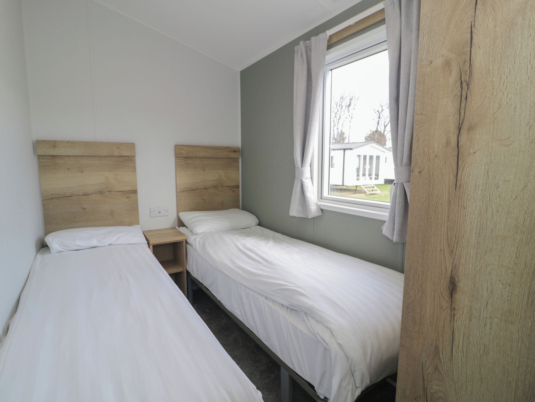 A small, clean room with two single beds, a wooden headboard, a nightstand, and a window with curtains.