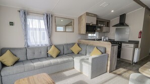 A modern living area with a grey sofa, a wooden coffee table, and a kitchenette with a stove and microwave. - Lido Gold 205 (Prestatyn)