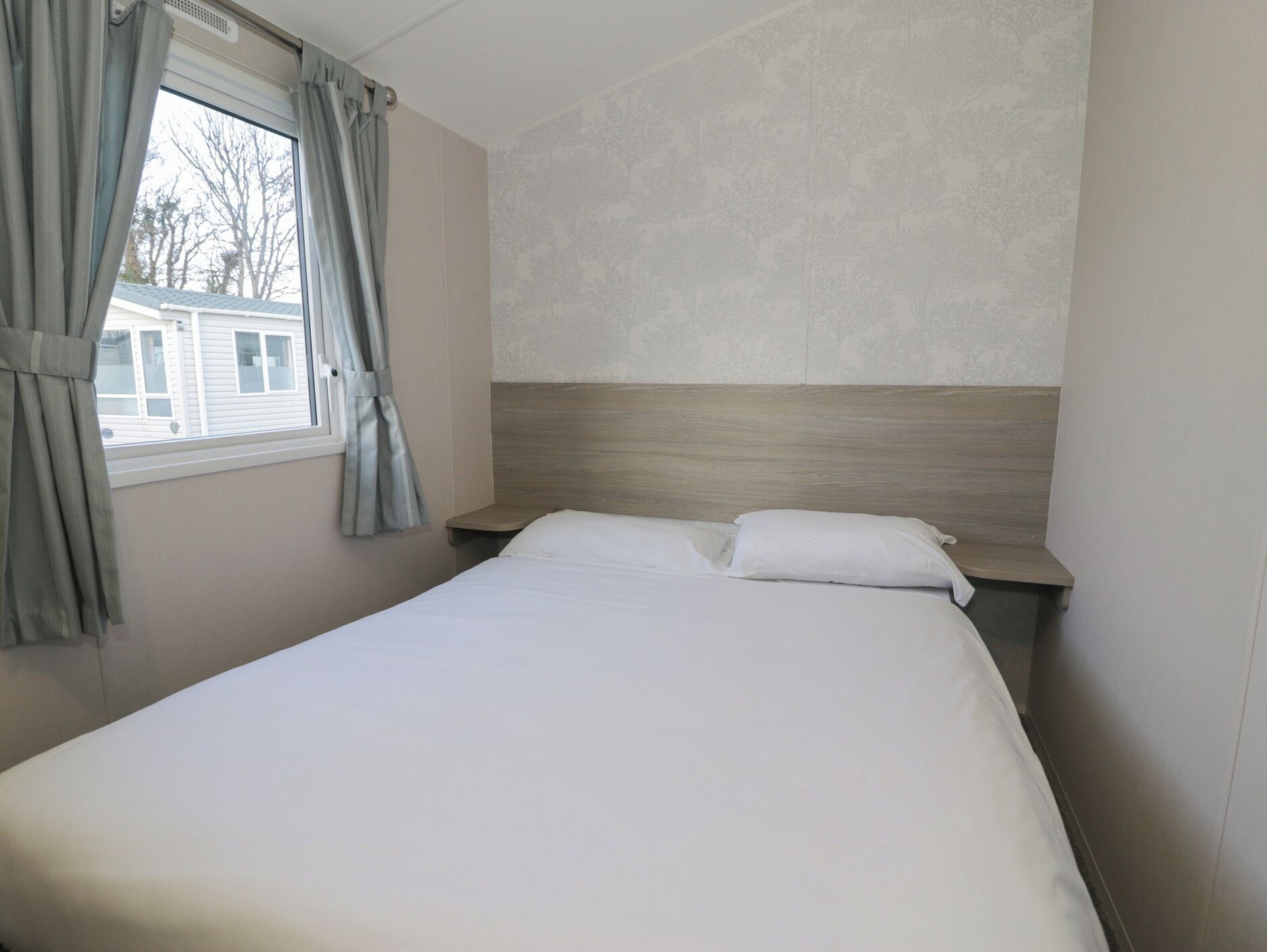 A hotel room with a bed, a nightstand, a window with curtains, and a wall with a subtle pattern.