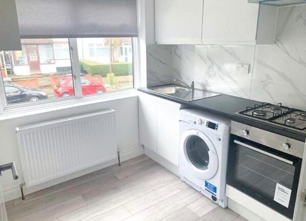 A compact kitchen with a built-in washer, a gas stove, and a window overlooking a residential street. - Sunny Gardens 3-bed Apartment in London (London)