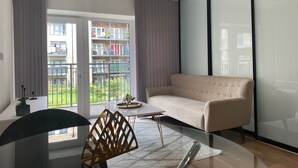 Ένα μοντέρνο σαλόνι με έναν καναπέ, ένα τραπεζάκι σαλονιού από γυαλί και θέα σε ένα μπαλκόνι με κάγκελο και φυτά.