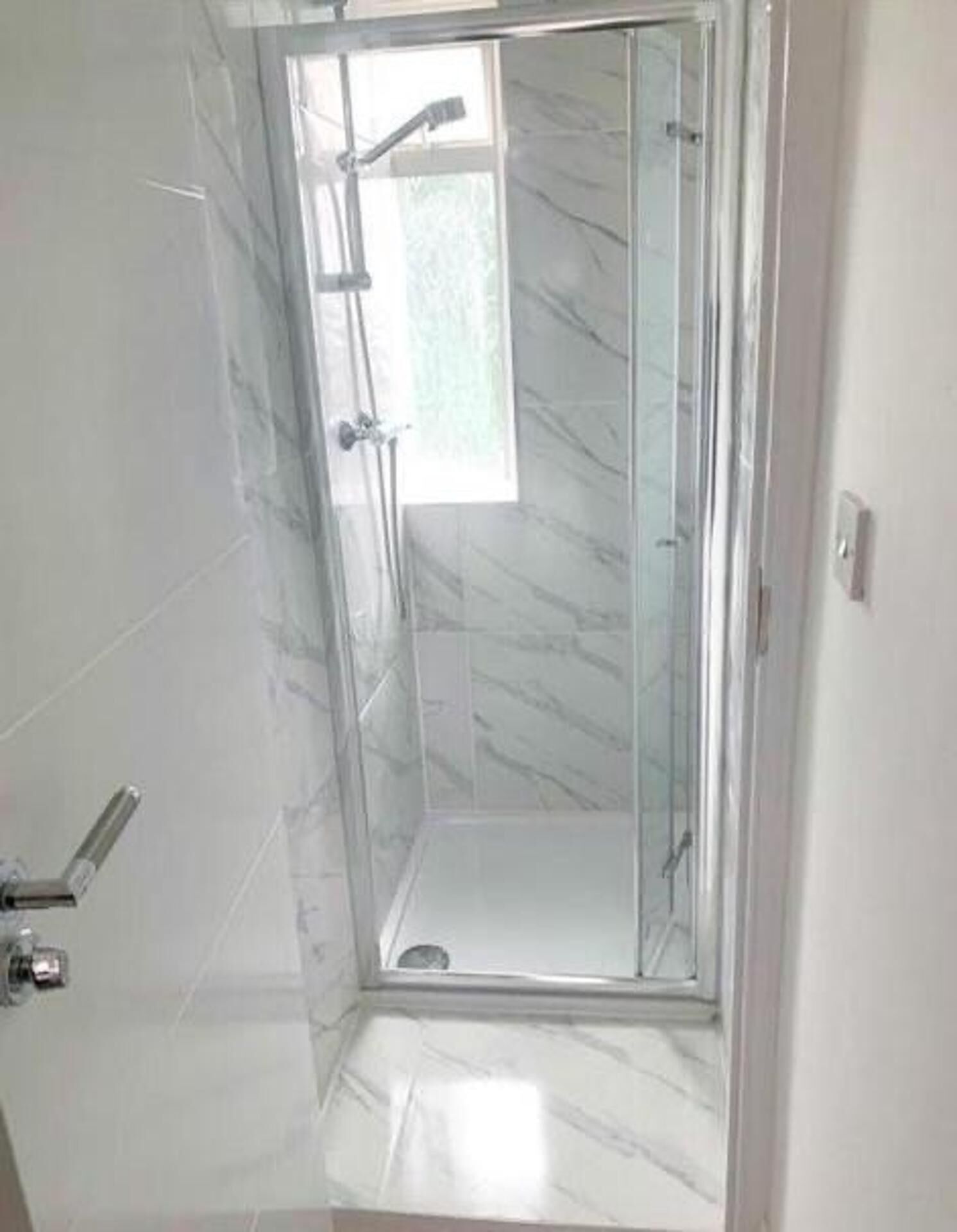 Ducha con cerramiento de vidrio, suelo de mármol y una ventana pequeña.