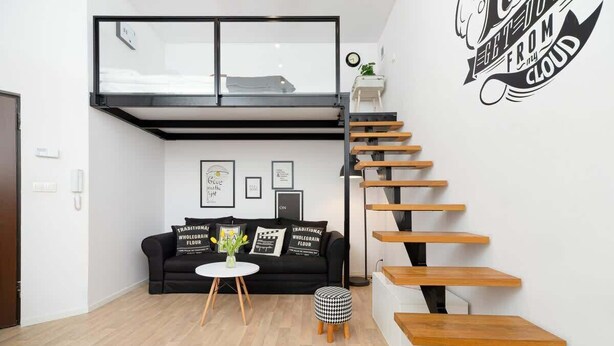 Un espacio habitable moderno con un sofá negro, una mesita redonda y un taburete a cuadros blanco y negro. Una escalera de madera con estructura metálica negra conduce a una zona superior.