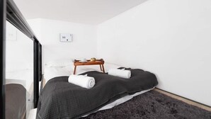 Habitación pequeña y moderna con una cama, una mesita de madera con una taza y un libro, y una alfombra mullida.