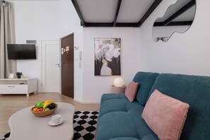 Un salon moderne avec un canapé de couleur turquoise, une table basse blanche ornée de fruits, un tapis noir et blanc, un téléviseur à écran plat et un tableau accroché au mur.