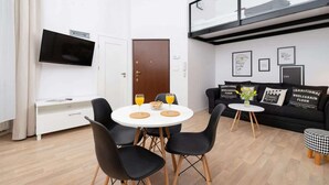 Una sala moderna con un sofá blanco, sillas negras, una mesa de comedor redonda con zumo de naranja, un televisor de pantalla plana y un techo estilo loft.