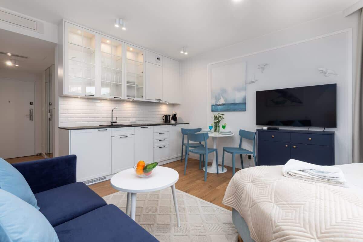 A modern hotel room with a kitchenette, a flat-screen TV, a sofa, and a dining area.