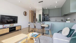 A modern living room with a flat-screen TV, a dining area with chairs and a table, and a kitchen with cabinets and appliances.