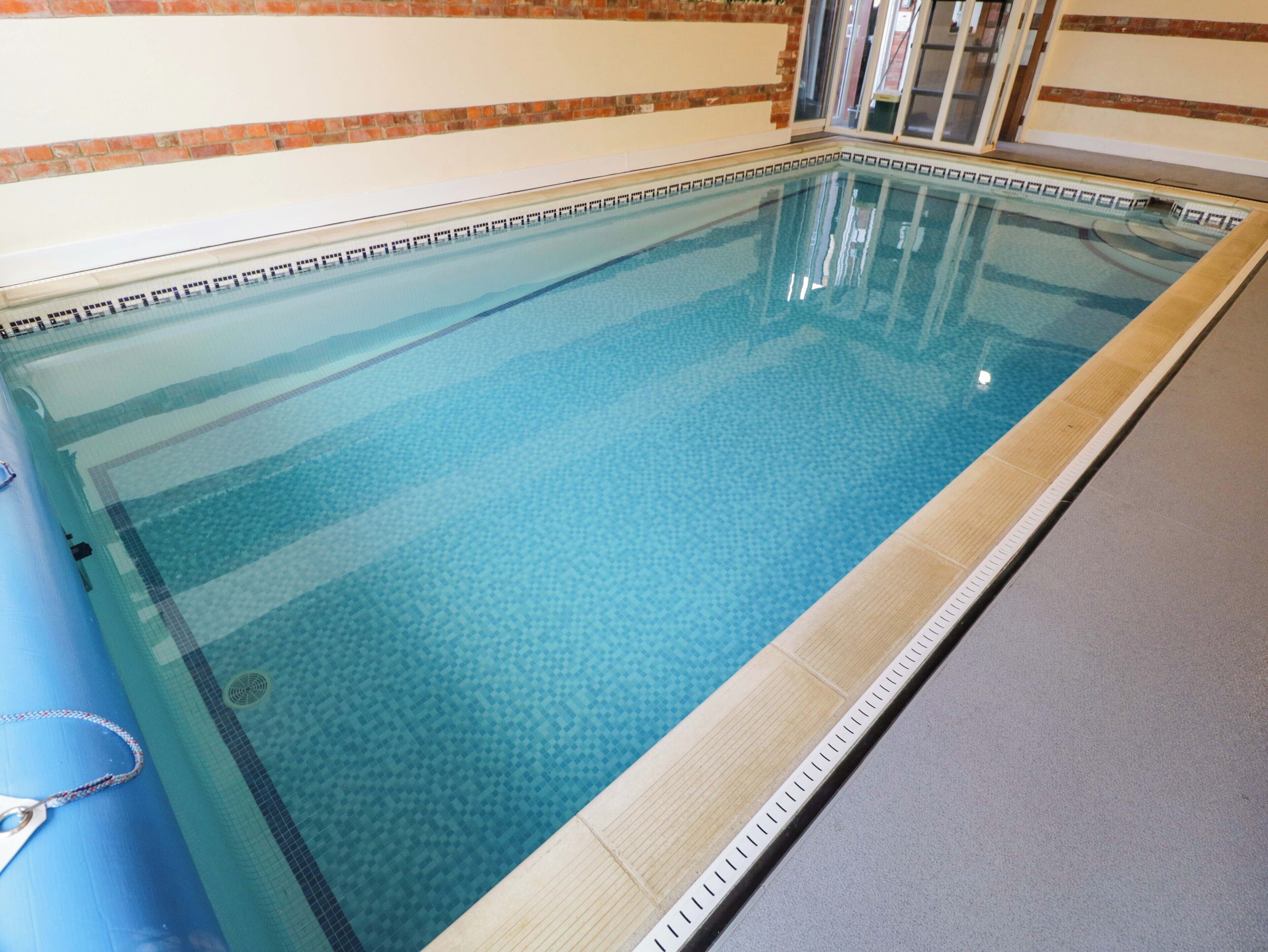 A rectangular indoor swimming pool with clear water, surrounded by a tiled deck and a brick wall.