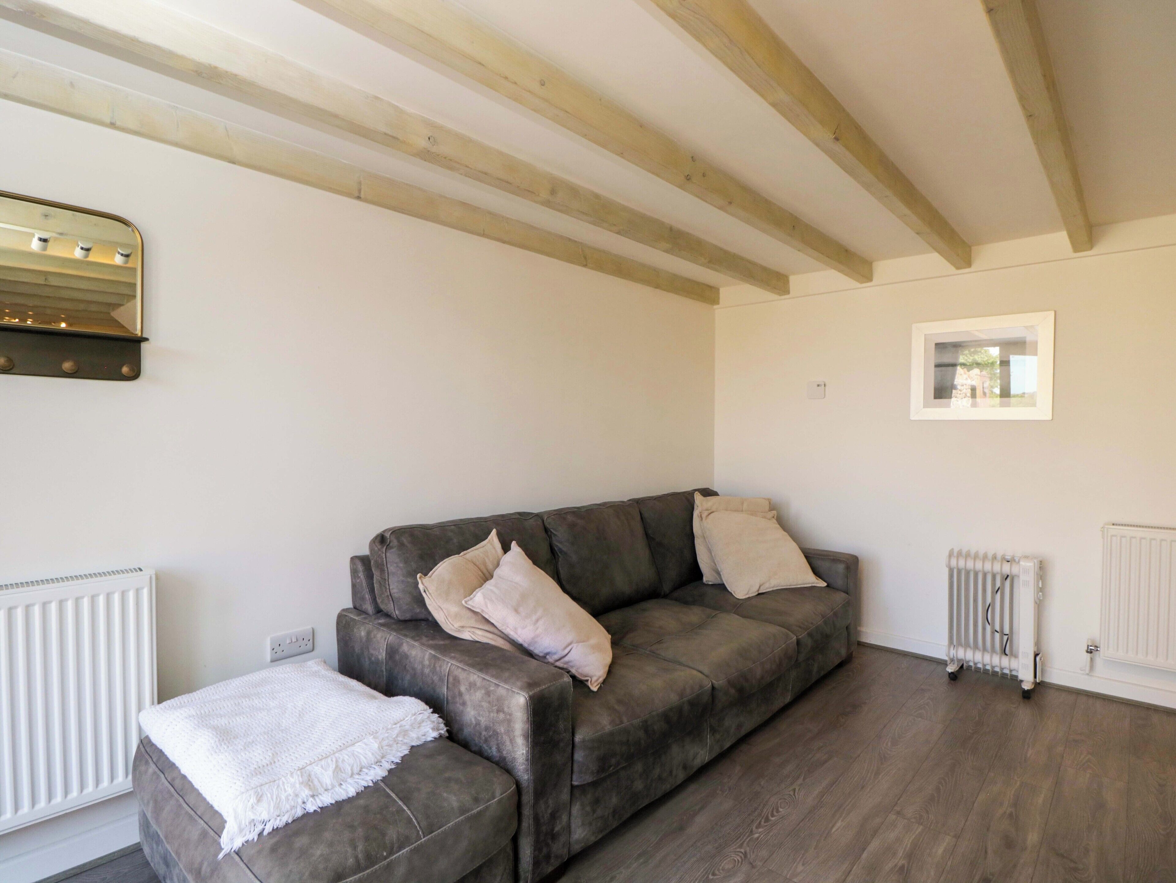 Ein Wohnzimmer mit grauem Sofa, weißer Decke, Holzbalken an der Decke, weißem Heizkörper und einem gerahmten Bild an der Wand.