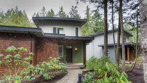 A modern two-story house with a brick facade and a green door, surrounded by a garden with various plants and a paved pathway.
