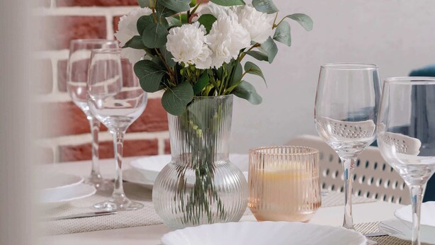 A dining table set with white plates, clear wine glasses, and a vase with white flowers.