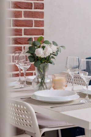 Una mesa de comedor dispuesta con platos blancos, copas de vino transparentes y un florero con flores blancas.