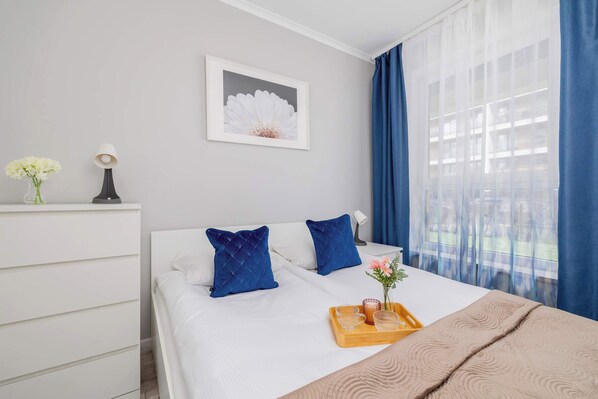 Habitación de hotel ordenada con una cama, una mesita de noche con lámpara, un cuadro en la pared y una ventana con cortinas azules.