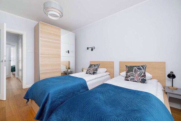 A hotel room with two beds, a wardrobe, a bedside table with a lamp, and a door leading to another room.