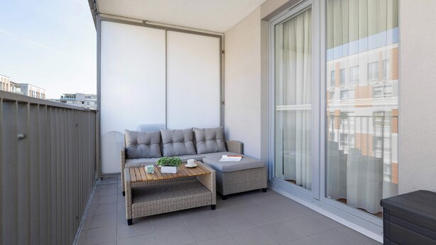 Ein Balkon mit einer grauen Couch, einem kleinen Tisch mit einer Pflanze und einem Fenster mit Vorhängen.
