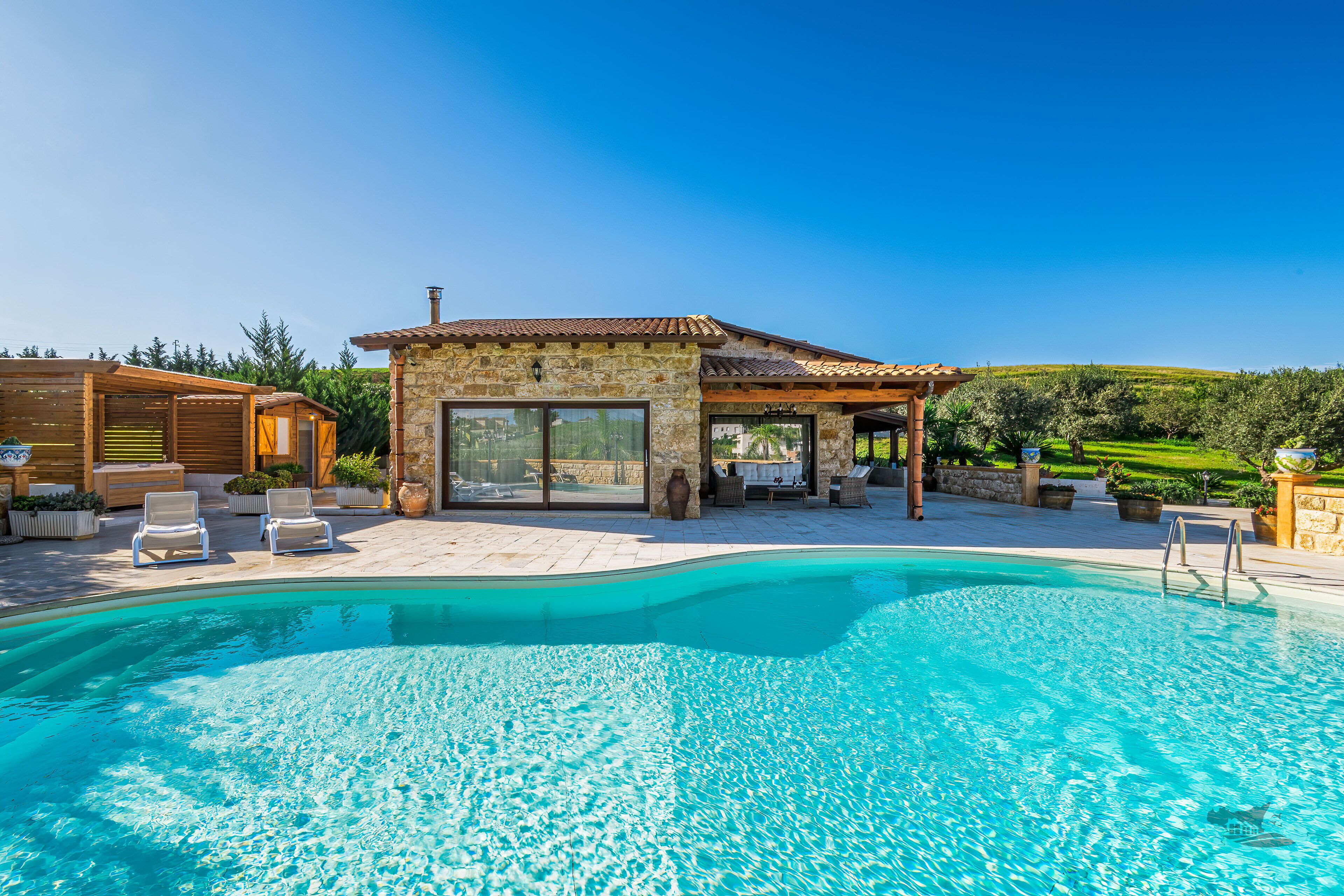 Un espace piscine avec un bâtiment en pierre, des chaises longues et un ciel d’un bleu éclatant.