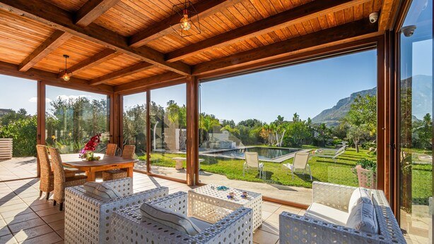 A covered outdoor seating area with wicker furniture, large windows, and a view of a landscaped garden and mountains. - Villa DellAlbero: Privacy, Relax and Unique Pool (Alcamo)