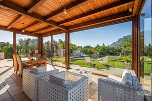 Un espacio al aire libre cubierto con mobiliario de mimbre, ventanas amplias y vistas a un jardín cuidado y montañas.