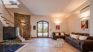 A spacious living room with a large TV, a stone wall, a staircase, and a glass coffee table. - Villa DellAlbero: Privacy, Relax and Unique Pool (Alcamo)