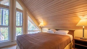 Un dormitorio con techo inclinado, vigas de madera, una cama con colcha marrón, dos lámparas de noche y ventanas amplias que dan a vista de árboles.