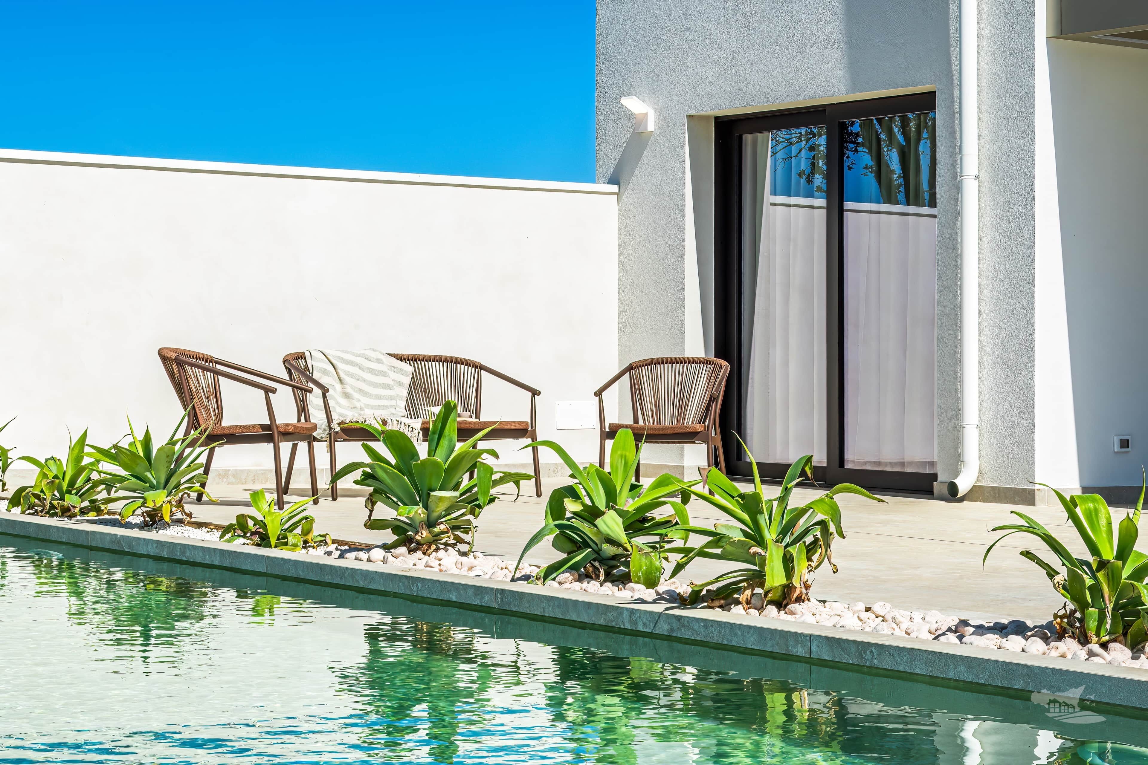 A pool area with chairs and plants, a clear sky, and a building with a door and window.