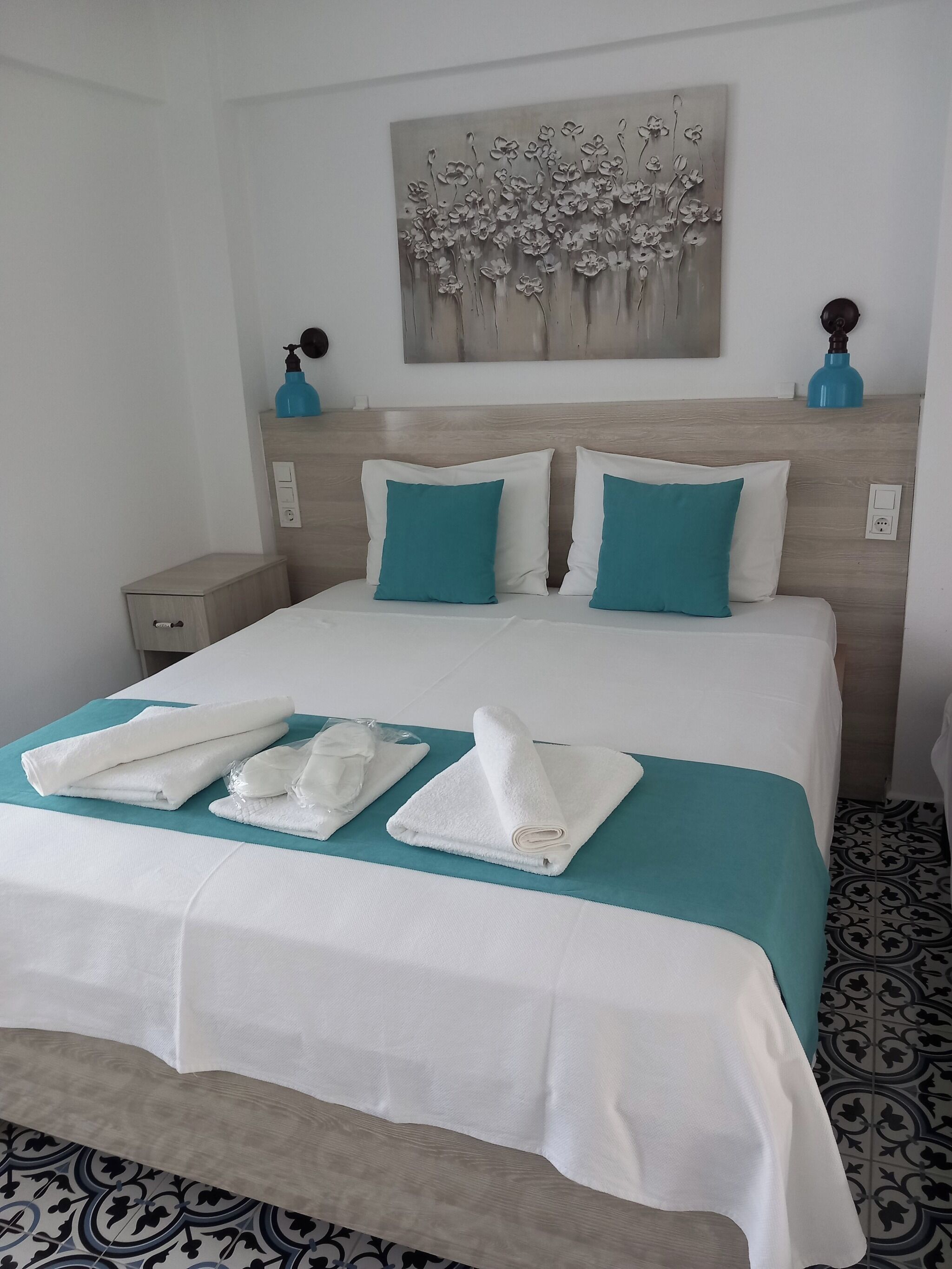 A neatly made bed with white and teal bedding, a wall art piece above, and a patterned floor.
