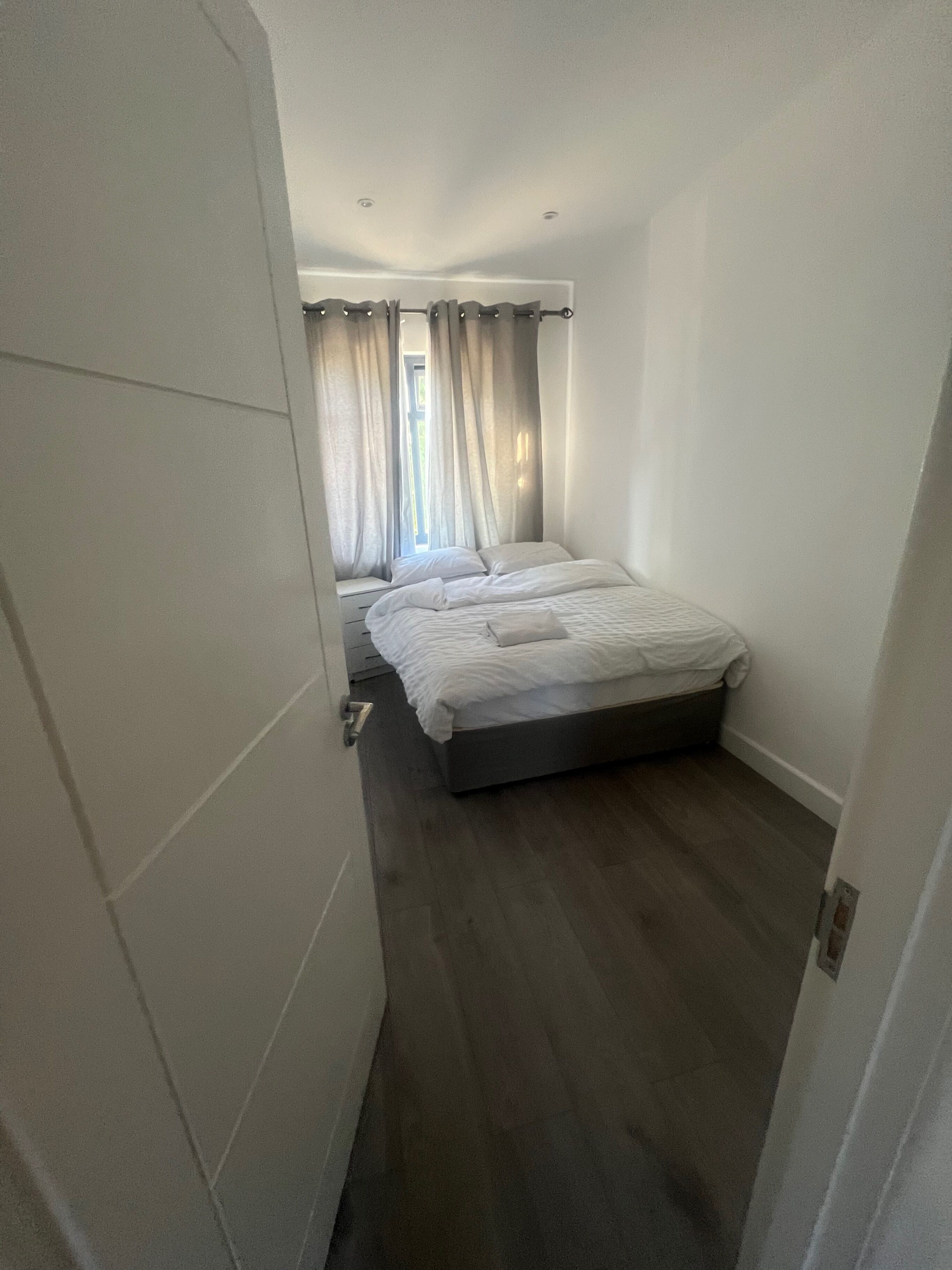 A narrow hotel room with a single bed, a small window with curtains, and a door on the left.