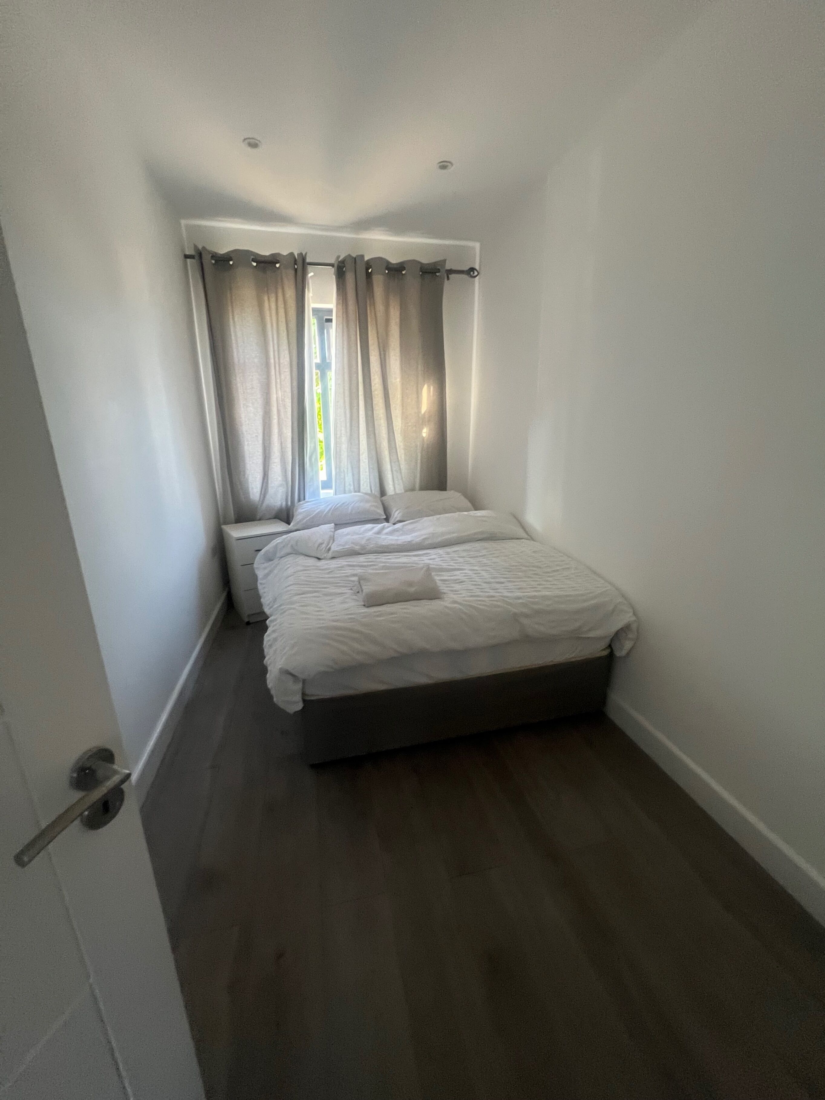 A small, narrow hotel room with a single bed, a window with curtains, and a door on one side.