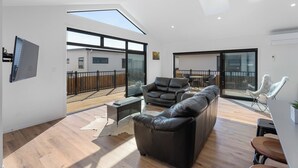 A modern living room with a large sofa, a coffee table, and a TV. There is a balcony with a view of a house.
