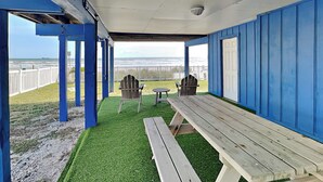 A covered outdoor seating area with a view of the beach, featuring a picnic table and two Adirondack chairs. - Bermuda Blue by Ryson Vacation Rentals (Galveston)