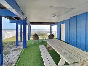 A covered outdoor seating area with a view of the beach, featuring a picnic table and two Adirondack chairs. - Bermuda Blue by Ryson Vacation Rentals (Galveston)