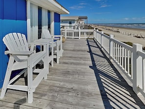 A wooden deck with white Adirondack chairs, a white fence, and a view of the beach and ocean. - Bermuda Blue by Ryson Vacation Rentals (Galveston)