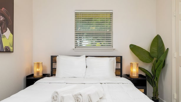 Een netjes opgemaakt bed met witte lakens en opgerolde handdoeken, een raam met zonwering, bedlampen en een grote groene plant.