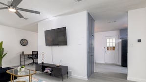 Een moderne woonkamer met een flatscreen televisie, een plafondventilator, een zwarte kantoorstoel, een klein bijzettafeltje en een kleurrijk vloerkleed.