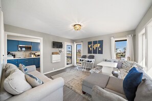 A modern living room with a sofa, a coffee table, and a dining area with a white table and chairs. There is a kitchen visible through an open door, and a large window provides a view of the outside. - Captain's Manor by Ryson Vacation Rentals (Galveston)