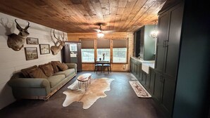 A rustic cabin interior with a wooden ceiling, a sofa, a dining table, and a kitchen area.