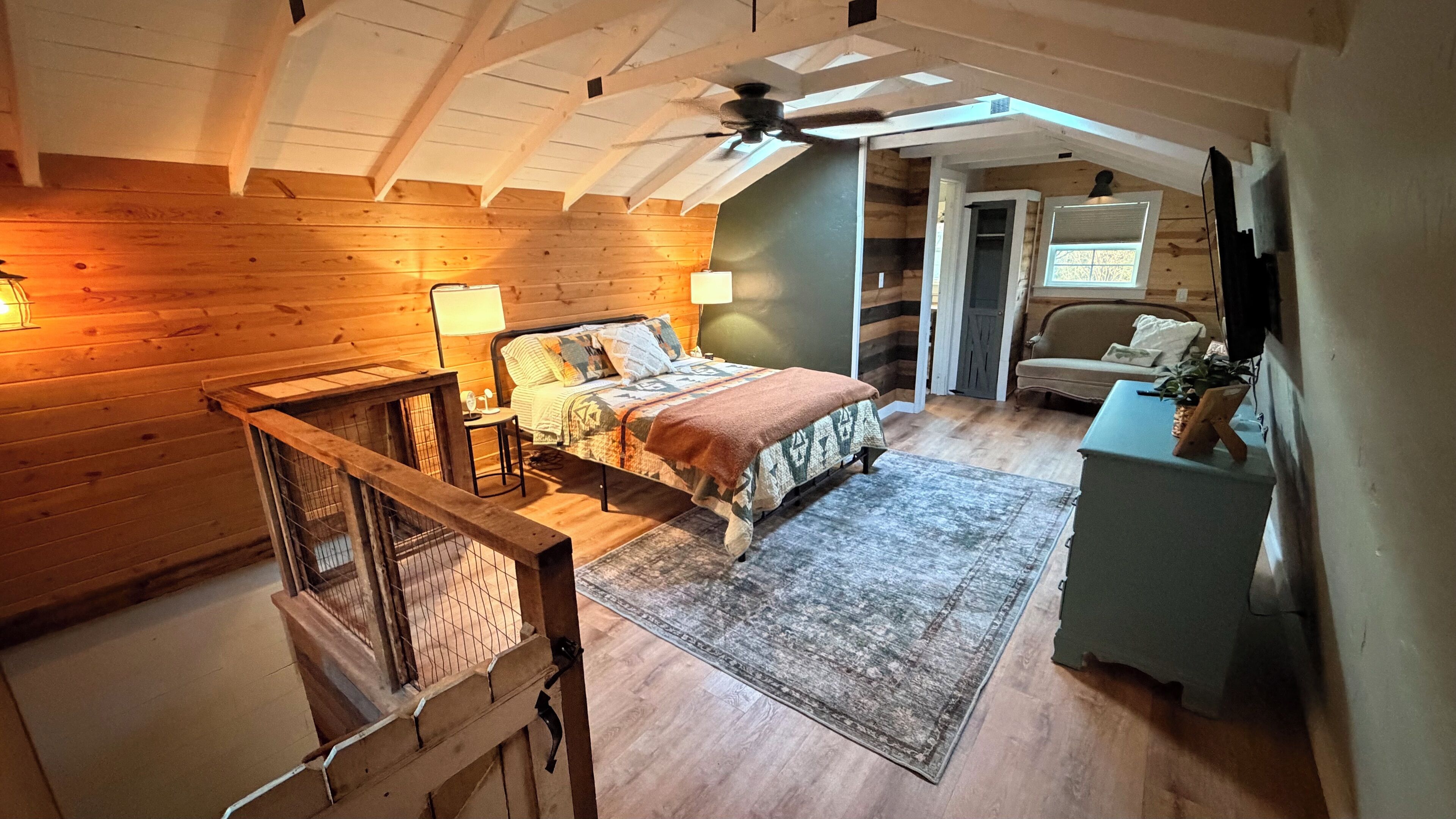 Une chambre avec un plafond en bois, un lit recouvert d’une couvre-lit à motifs, une table de chevet, une lampe et une vue sur un coin repas.