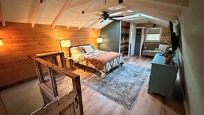 A bedroom with a wooden ceiling, a bed with a patterned bedspread, a nightstand, a lamp, and a view of a dining area. - Pond-View Cabin on 30 Acres / Dock + Fire Pit + Wildlife / Sleeps 4 (Emory)