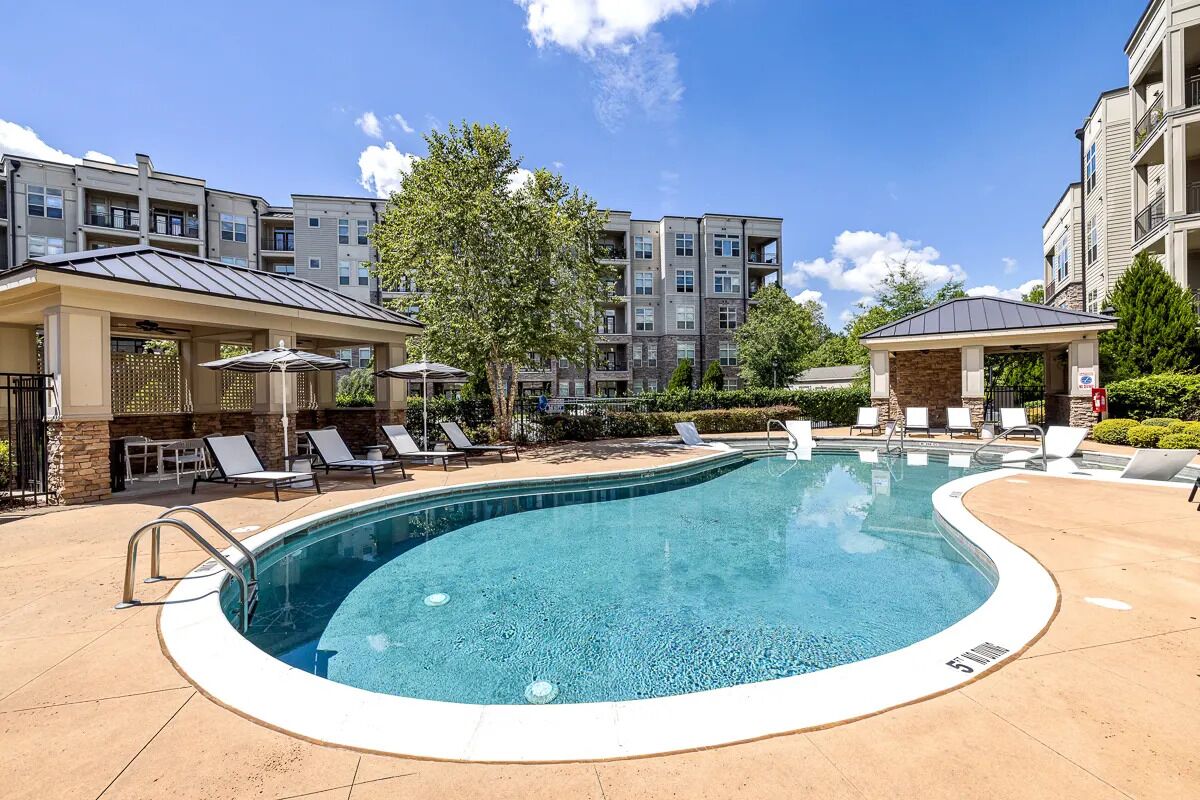 A swimming pool with lounge chairs and a covered area, surrounded by apartment buildings.