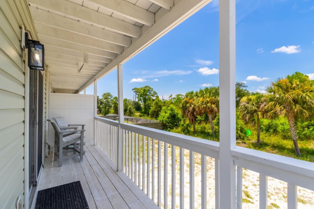 A covered porch with a bench and a door, overlooking a beach with palm trees and a clear sky.