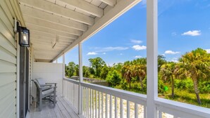 A covered porch with a bench and a door, overlooking a beach with palm trees and a clear sky.