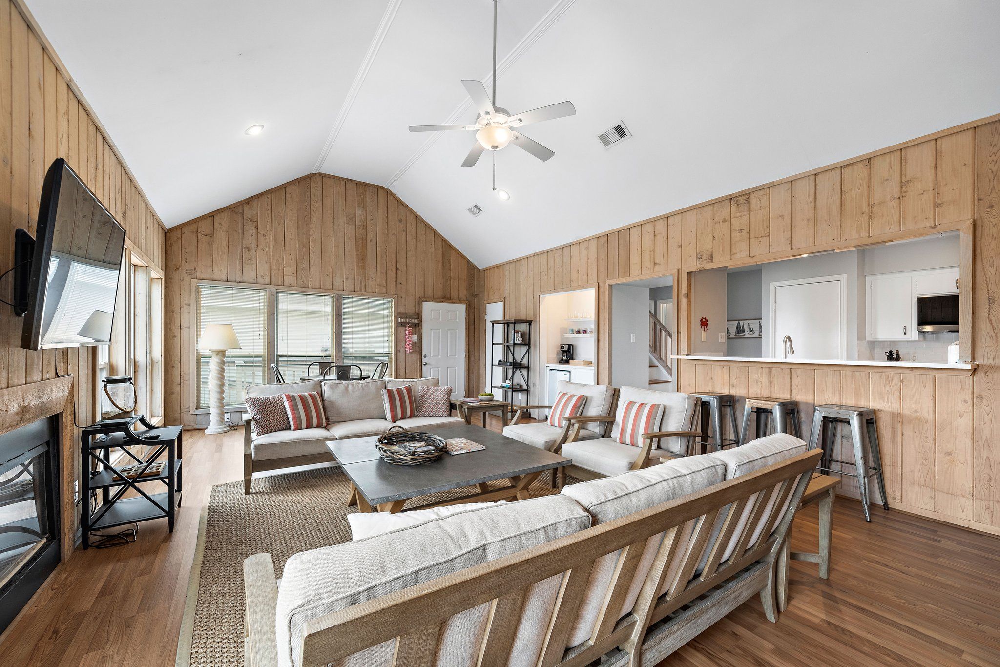 A spacious living area with a high ceiling, wooden paneling, a central seating area with sofas and a coffee table, a television mounted on the wall, and a kitchen area with a countertop and cabinets.