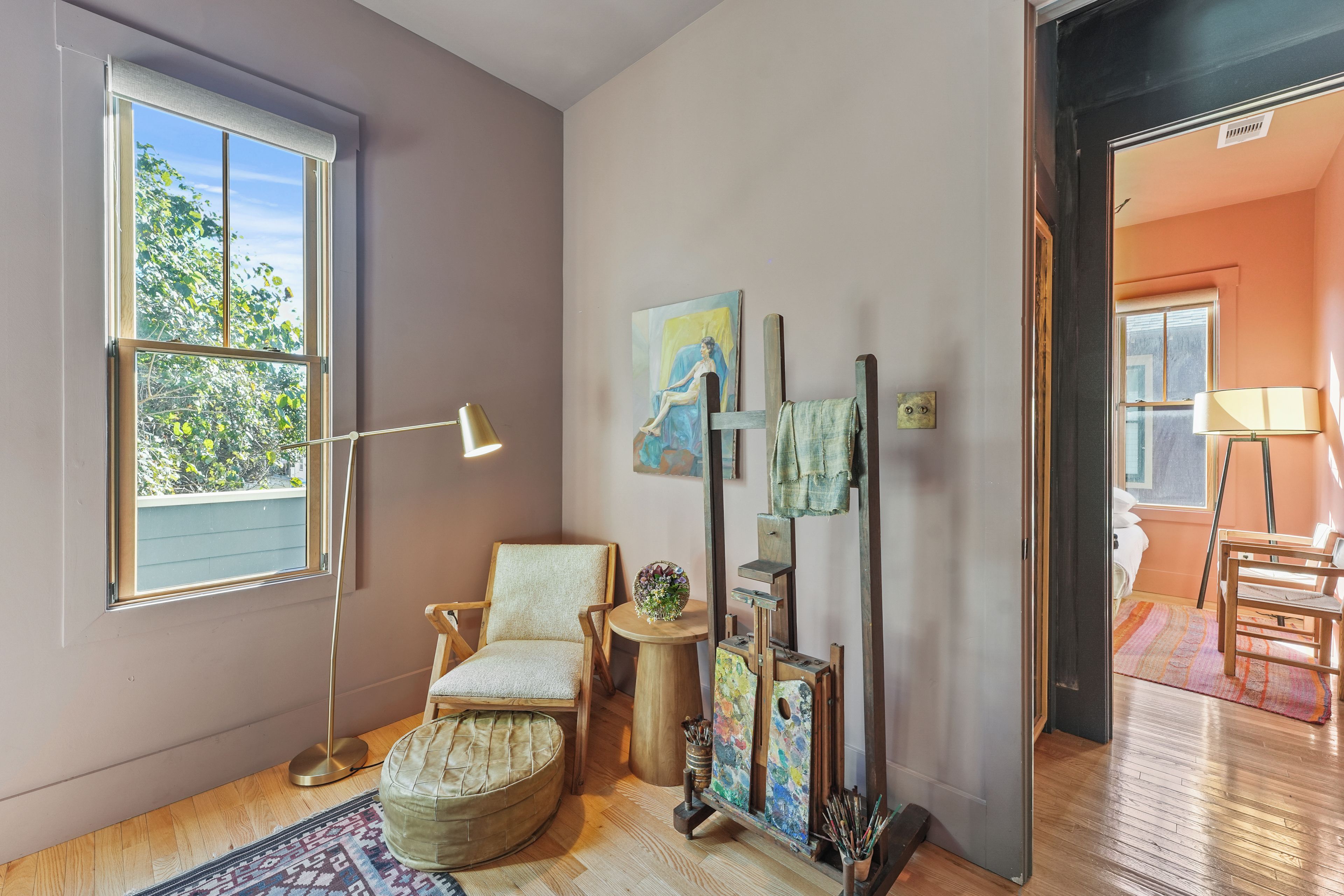 Ein Zimmer mit einem großen Fenster, einem Sessel, einer Stehlampe, einem kleinen Tisch mit einer Vase und einem Gemälde an der Wand.