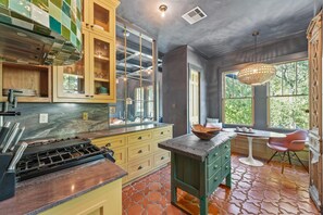 A kitchen with a gas stove, wooden cabinets, a glass-fronted pantry, and a dining area with a table and chair. - Ebb and Glow Retreat by Ryson Vacation Rentals (Galveston)