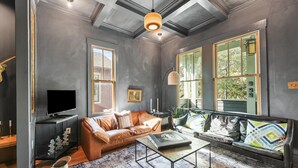 Un salon moderne avec un canapé en cuir, une table basse et un téléviseur posé sur un meuble noir. De grandes fenêtres laissent entrer la lumière naturelle et offrent une vue sur l’extérieur.