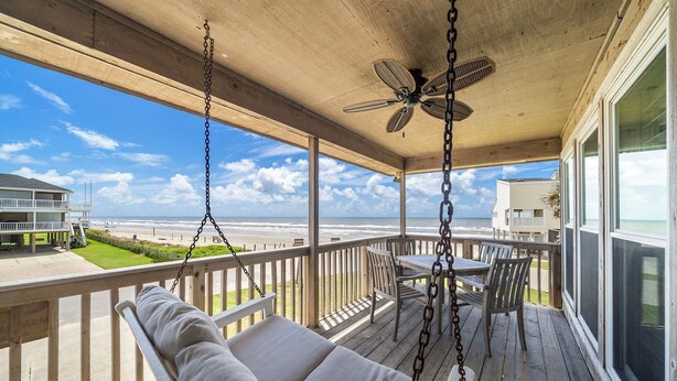 Une terrasse en bois avec un fauteuil suspendu et une table, offrant une vue sur une plage et l’océan.